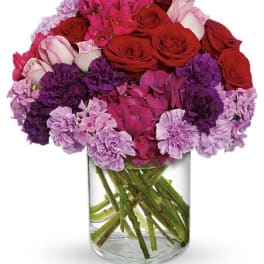Round arrangement of red and pink roses with purple and lavender carnations in a clear glass vase