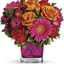 Bright arrangement of orange roses and pink gerbera daisies in a pink glass cube vase