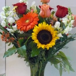 Mixed bouquet of roses, gerbera daisies, and sunflowers in a glass vase