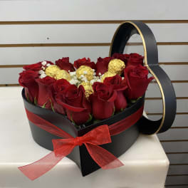 Heart-shaped black box of red roses with gold-wrapped chocolates and a red ribbon bow