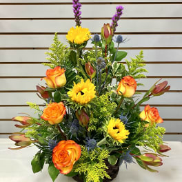 Basket arrangement with orange roses, yellow sunflowers, and purple accents