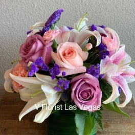 Compact arrangement of pink roses, lilies, and purple flowers in a green glass cube vase