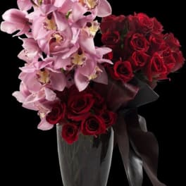 Pink orchids and red roses in a clear vase with a dark ribbon