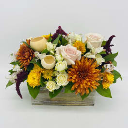 Low rectangular arrangement with orange and yellow mums, pale roses, and white blooms in a wooden box with decorative pods.
