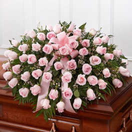Large spray of light pink roses with ribbon displayed on a wooden casket.