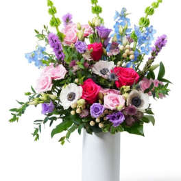 Tall mixed bouquet of pink roses, white anemones, and purple and blue blooms in a white vase