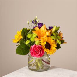 Mixed bouquet of sunflowers, roses, lilies, and purple blooms in a glass vase