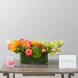Rectangular floral arrangement with coral roses, orange blooms, and green orchids in a glass vase