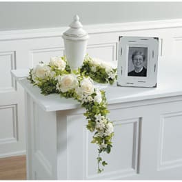 White funeral urn surrounded by white roses and greenery beside a framed portrait