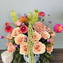 Peach roses and pink blooms arranged in a striped vase with a candle beside it