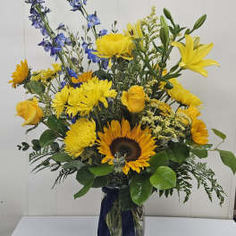 Tall bouquet of yellow and blue flowers in a glass vase with a dark ribbon