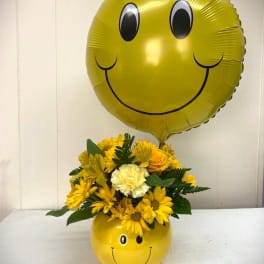 Yellow smiley-face balloon above a yellow flower arrangement in a matching container