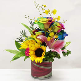 Bright mixed arrangement with rainbow roses, sunflower, pink lily and yellow orchids in a red glass vase