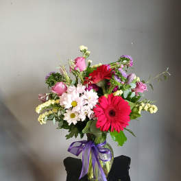 Mixed bouquet of pink, red, white, and purple flowers in a glass vase with a purple ribbon.