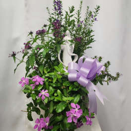 Purple flowers and greenery arranged around a white figurine with a lavender ribbon bow.