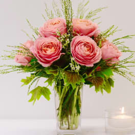 Pink roses arranged in a clear glass vase with greenery and a lit candle nearby