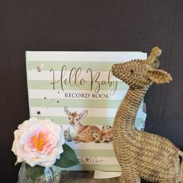 Single pink-and-white flower in a small glass vase beside a baby record book and woven deer figurine.