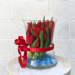 Red tulips in a clear glass vase with a red ribbon and blue stones