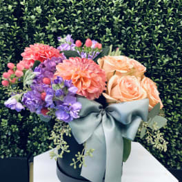 Bouquet of peach roses, purple blooms, and coral dahlias in a black hat box with a blue ribbon.