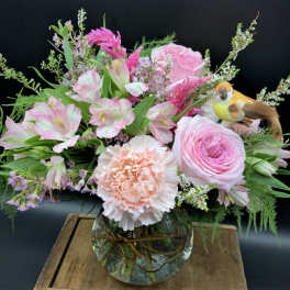 Pink roses, carnations, and mixed flowers in a round glass vase with a small decorative bird pick