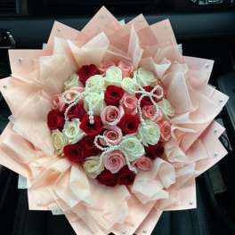 Round bouquet of red, pink, and white roses wrapped in layered peach paper with pearl accents.
