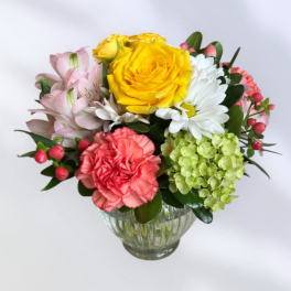 Mixed bouquet of yellow roses, pink flowers, white daisies, and green hydrangea in a glass vase