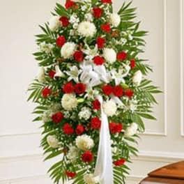 Tall standing spray of red and white flowers with a white bow on an easel beside a wooden casket.