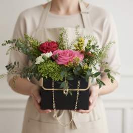 Mixed pink and red flowers in a black box with a gold chain handle