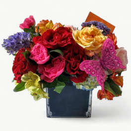 Bright mixed bouquet of roses and other blooms in a square glass vase with a pink butterfly pick
