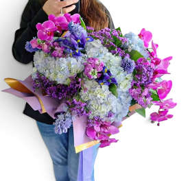 Large bouquet of purple orchids, hydrangeas, and mixed blooms wrapped in lavender paper