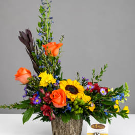 Mixed floral arrangement with orange roses, sunflowers, and blue accents in a textured container