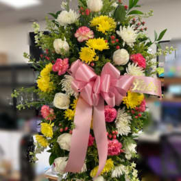 Standing spray of pink, yellow, and white flowers with a large pink bow on an easel.