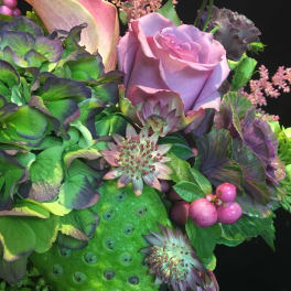 Arrangement of lavender rose, calla lily, hydrangea, berries and lotus pod in rich green and purple tones.