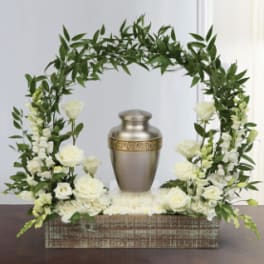 White roses and greenery arranged around a metal urn in a wooden box