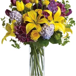 Bouquet of yellow lilies, purple flowers, and roses in a clear glass vase