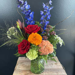 Mixed bouquet with red roses, blue delphinium, and orange blooms in a green glass vase