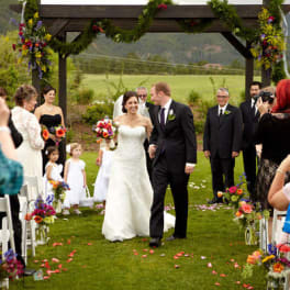 Bride and groom walking down an outdoor wedding aisle with floral arch and guests