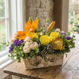 Mixed bouquet in a rustic rectangular container with yellow, orange, purple, and cream flowers