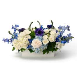 Low centerpiece of white and blue flowers, including roses and hydrangeas, in a rectangular white container
