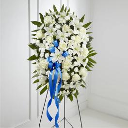 White funeral spray with roses and lilies on a stand, accented by a blue ribbon