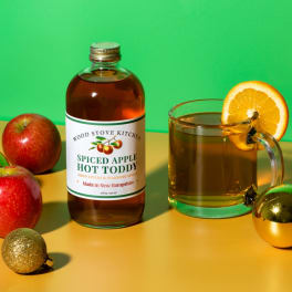 Bottle of spiced apple hot toddy mix beside a mug with orange slice and apples