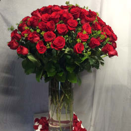 Large arrangement of long-stemmed red roses in a tall clear glass vase with petals scattered below.