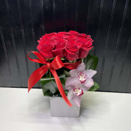 Compact arrangement of red roses with pink orchids in a square white vase with a red ribbon bow