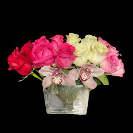 Low arrangement of red, pink, and ivory roses with two pink cymbidium orchids in a clear glass cube vase.
