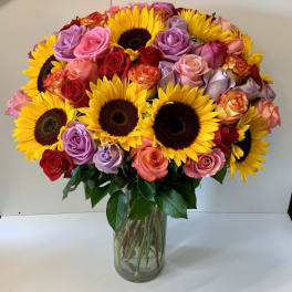 Tall glass vase of yellow sunflowers mixed with red, pink, and lavender roses