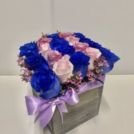 Square wooden box of bright blue and light pink roses with tiny pink blooms and a lavender bow.