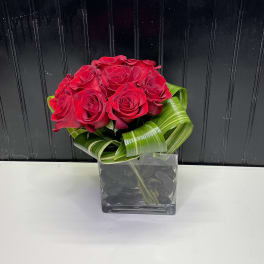 Compact arrangement of red roses with looped green foliage in a square glass vase with black stones