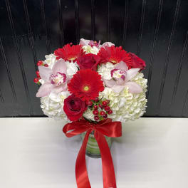 Red gerbera daisies, roses, pink orchids and white hydrangeas in a glass vase with red ribbon