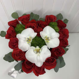Heart-shaped arrangement of red roses with white orchids and green moss accents.