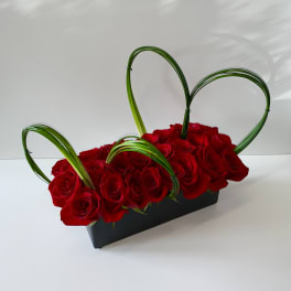 Rectangular arrangement of red roses in a black container with looped green blades forming hearts.